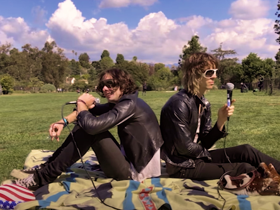 The two members of HELLP sitting on a picnic blanket in a park holding microphones.